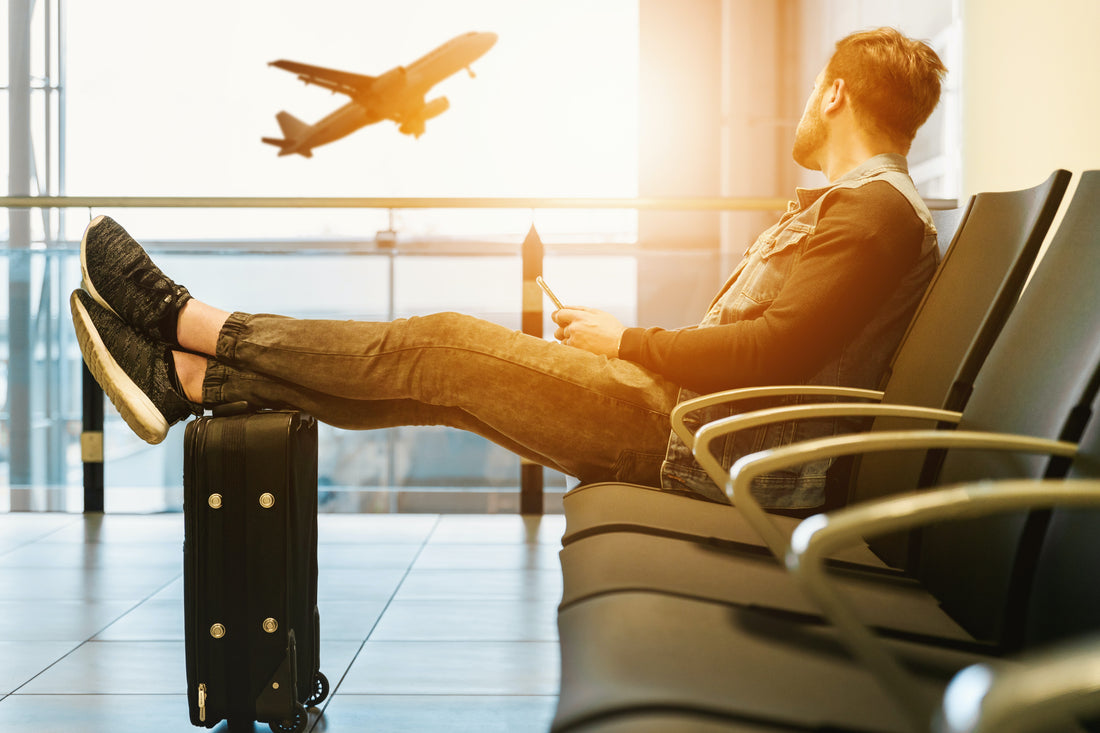 Guy sitting at the airport with his legs resting on his suitcase as he watches a plane take off outside the window